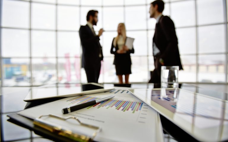 Technological devices and financial document at workplace on background of three business partners discussing ideas