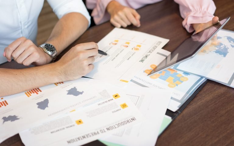 Hands of businesspeople discussing marketing reports and presentation on tablet. Close-up of young Caucasian businessman and businesswoman sitting at table and working over documents. Teamwork concept
